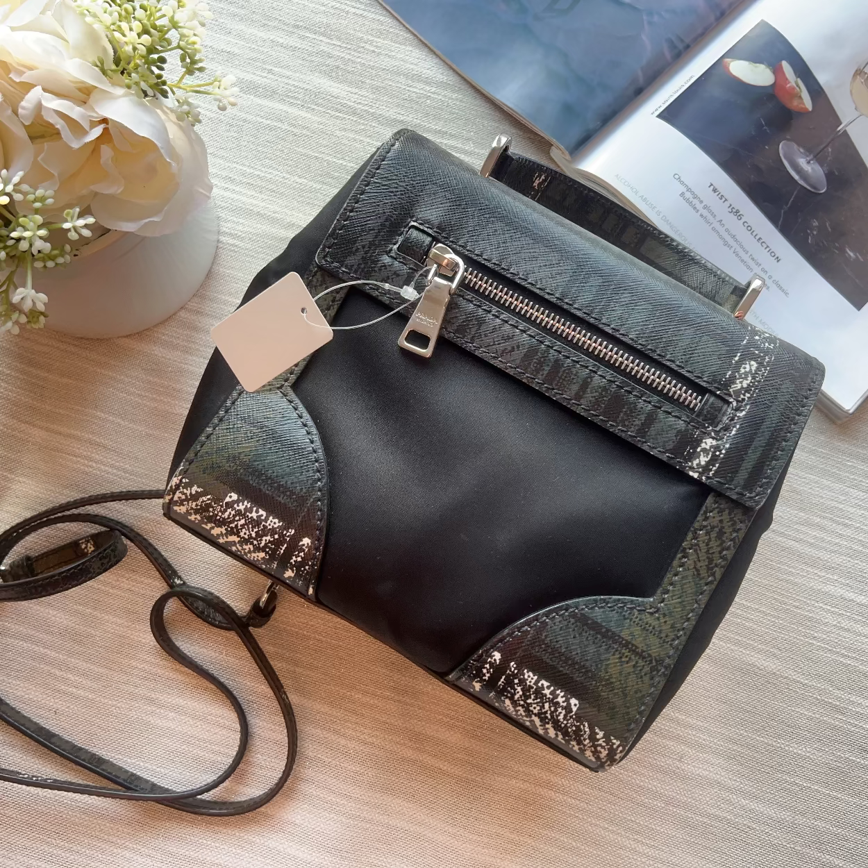 Black handbag with a strap on a light wooden surface, next to a vase of flowers and a magazine.