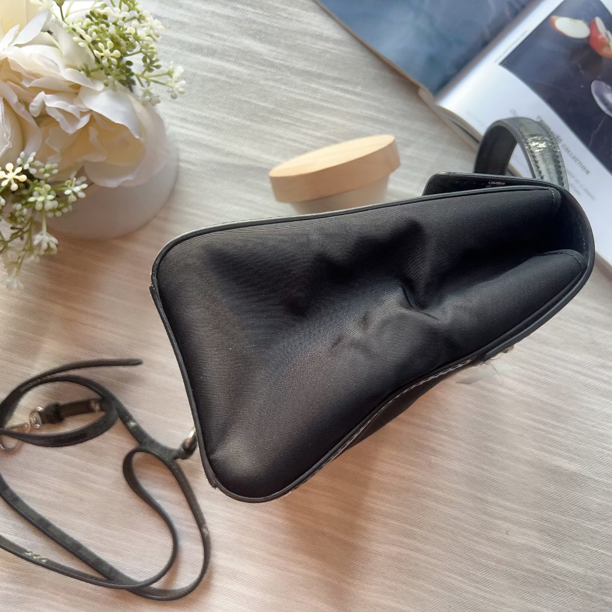 Black sunglasses case on a light wooden surface with a vase of flowers and a book in the background.