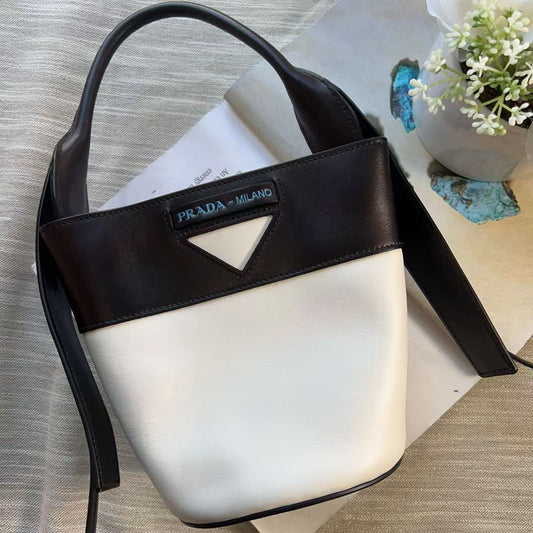 An Emporio Armani shoulder bag in white and dark brown leather, styled on a textured gray fabric surface with a book and small flowers in the background.