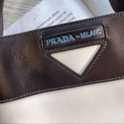 Close-up of a dark brown leather Prada bag strap with a triangular cutout and a vertical Prada Milano logo patch, resting on a light-colored surface.