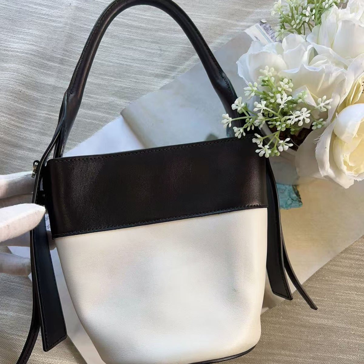 A color-blocked leather handbag with a white front panel, a wide dark brown band, and dark brown straps, styled on a beige textured fabric with white flowers in the background.