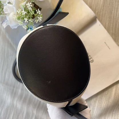 A top-down view of a round black leather bag or pouch with white trim, resting on a light-colored textured surface next to an open book and white flowers.