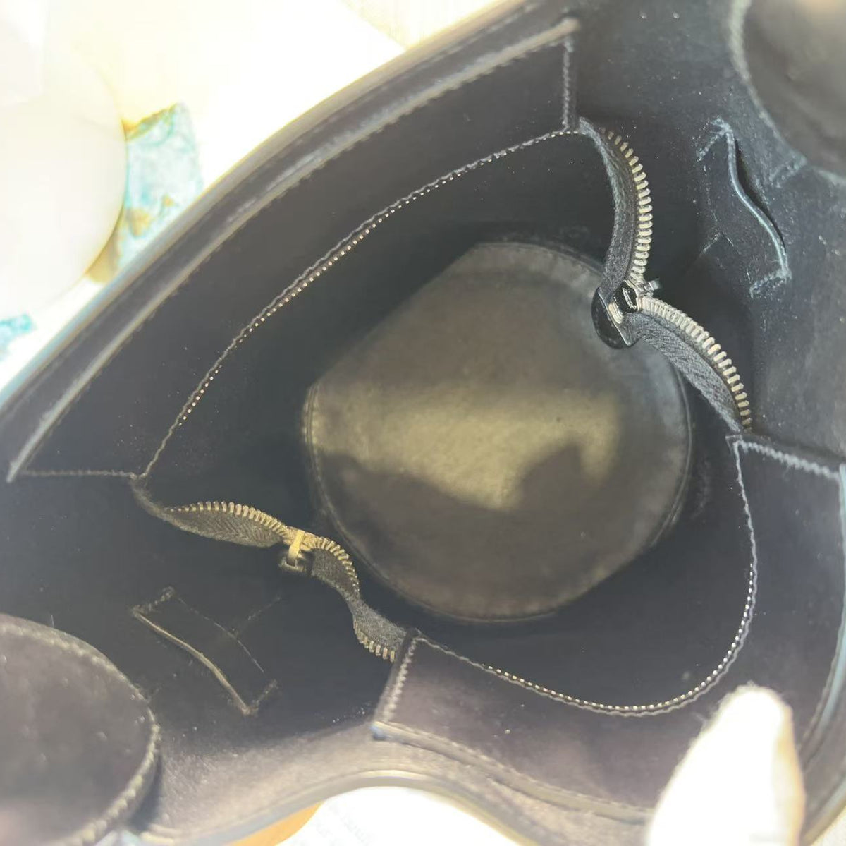 Close-up view from inside a black leather bag, showing a circular black insert or pouch with silver-toned zipper trim and interior stitching.