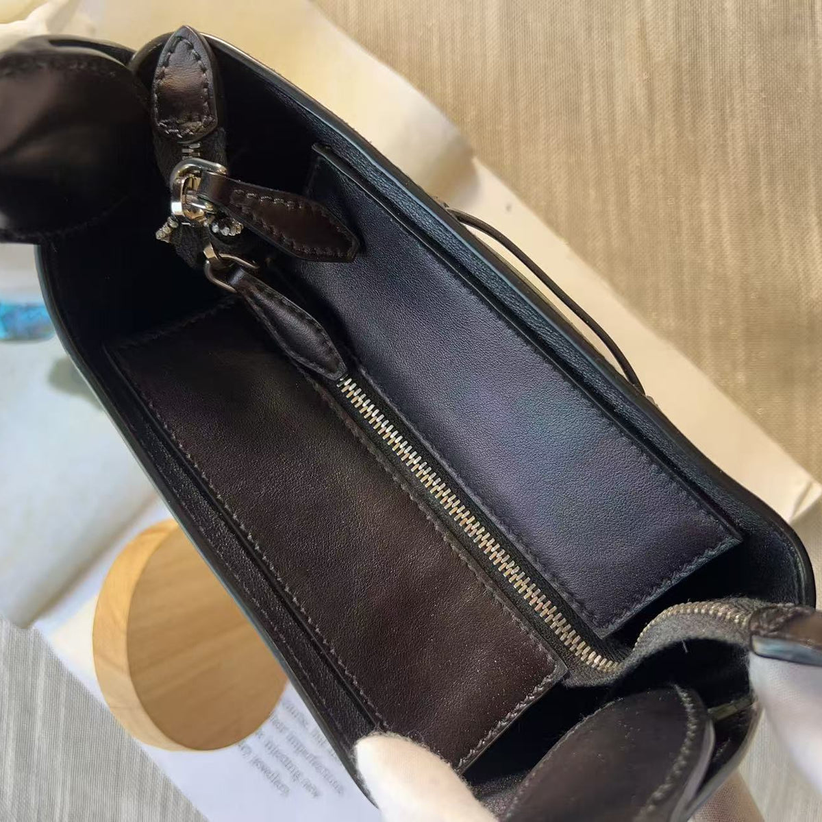 A close-up of a dark brown leather handbag interior, showing a zippered compartment, stitched lining, and a small leather pull tab, photographed on a light wood surface with a white box in the background.