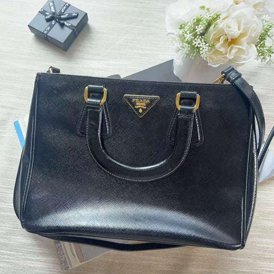 A black Prada handbag with gold-tone hardware and the iconic triangular Prada logo plaque on the front, displayed on a light wood surface with decorative props including a small gift box and white flowers.