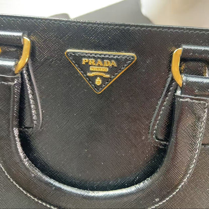 Close-up of a black Prada handbag featuring the signature gold-tone triangular Prada logo plaque on a textured leather surface.