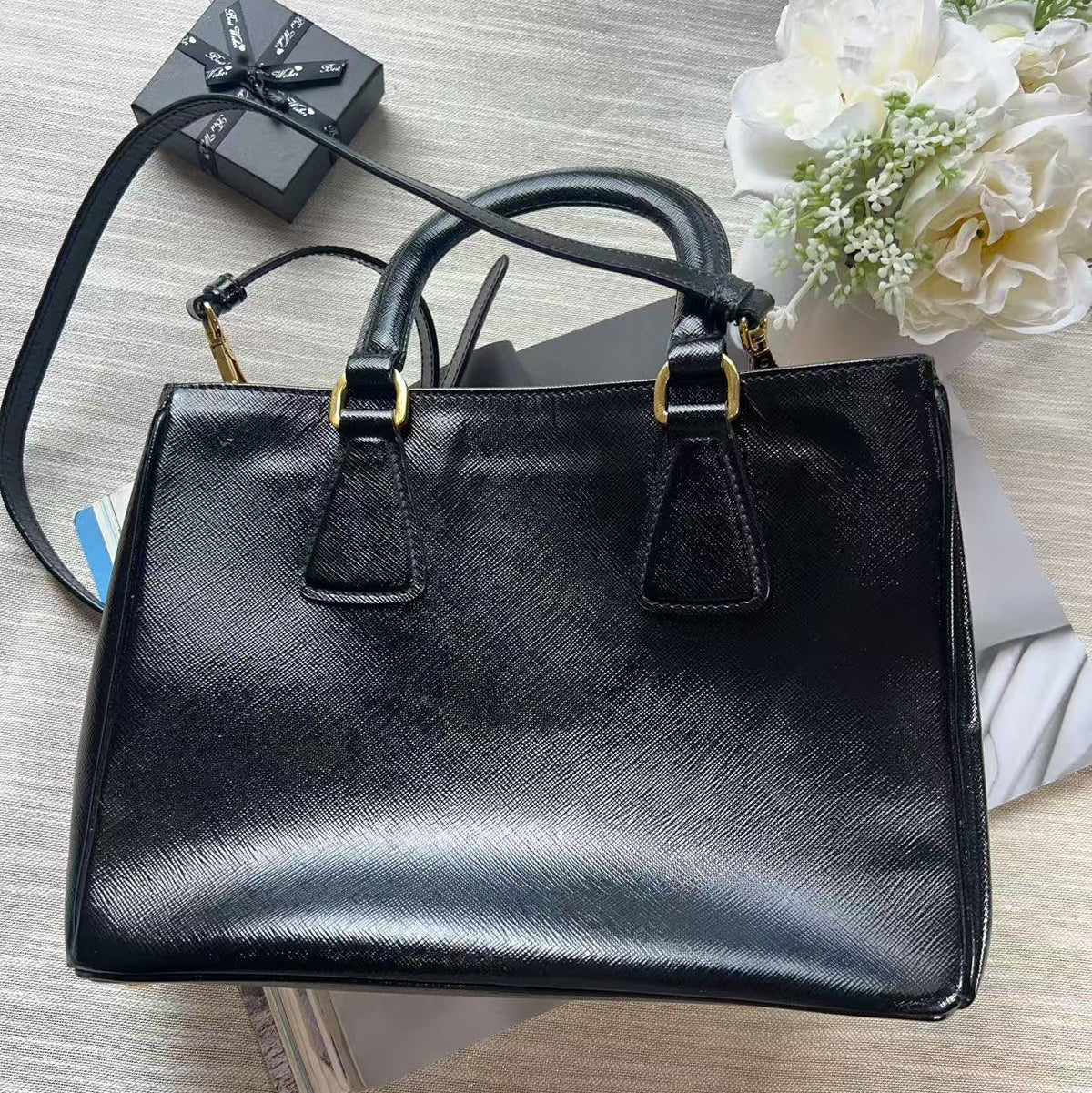 A black textured leather handbag with gold-tone hardware, displayed on a light wood surface with a small black gift box, white flowers, and a folded white garment as styling props.