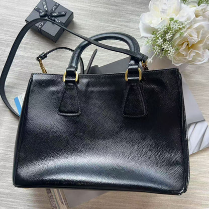 A black textured leather handbag with gold-tone hardware, displayed on a light wood surface with a small black gift box, white flowers, and a folded white garment as styling props.