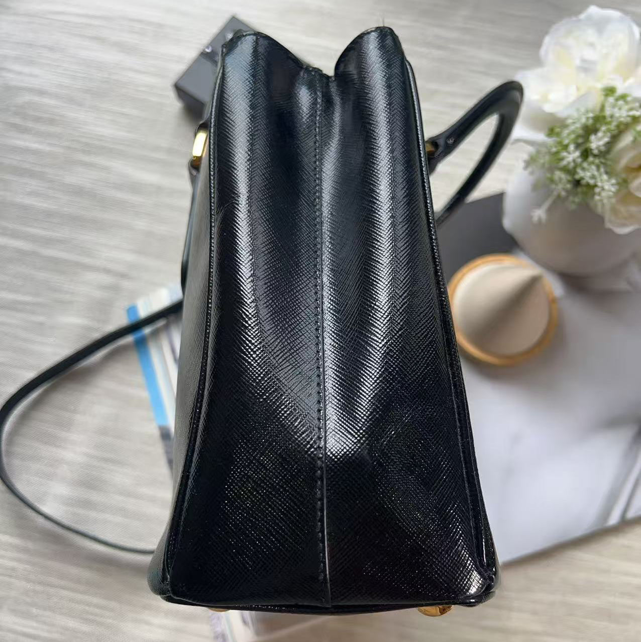 A black textured leather handbag shown from a top-down angle on a light wood surface, with a small vase of flowers and papers in the background.