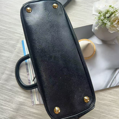 A black textured leather handbag shown from the bottom, featuring gold-tone metal studs at the corners and a black handle, photographed on a light wood surface with decorative props.