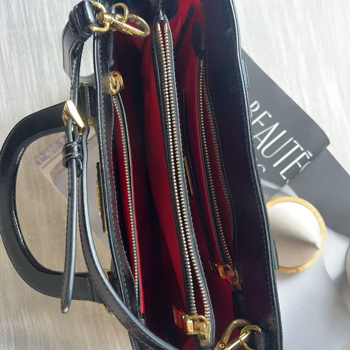 Black leather handbag shown open from above, revealing a bright red interior lining with multiple zip compartments and gold-tone hardware, placed on a light wood surface with a magazine underneath.
