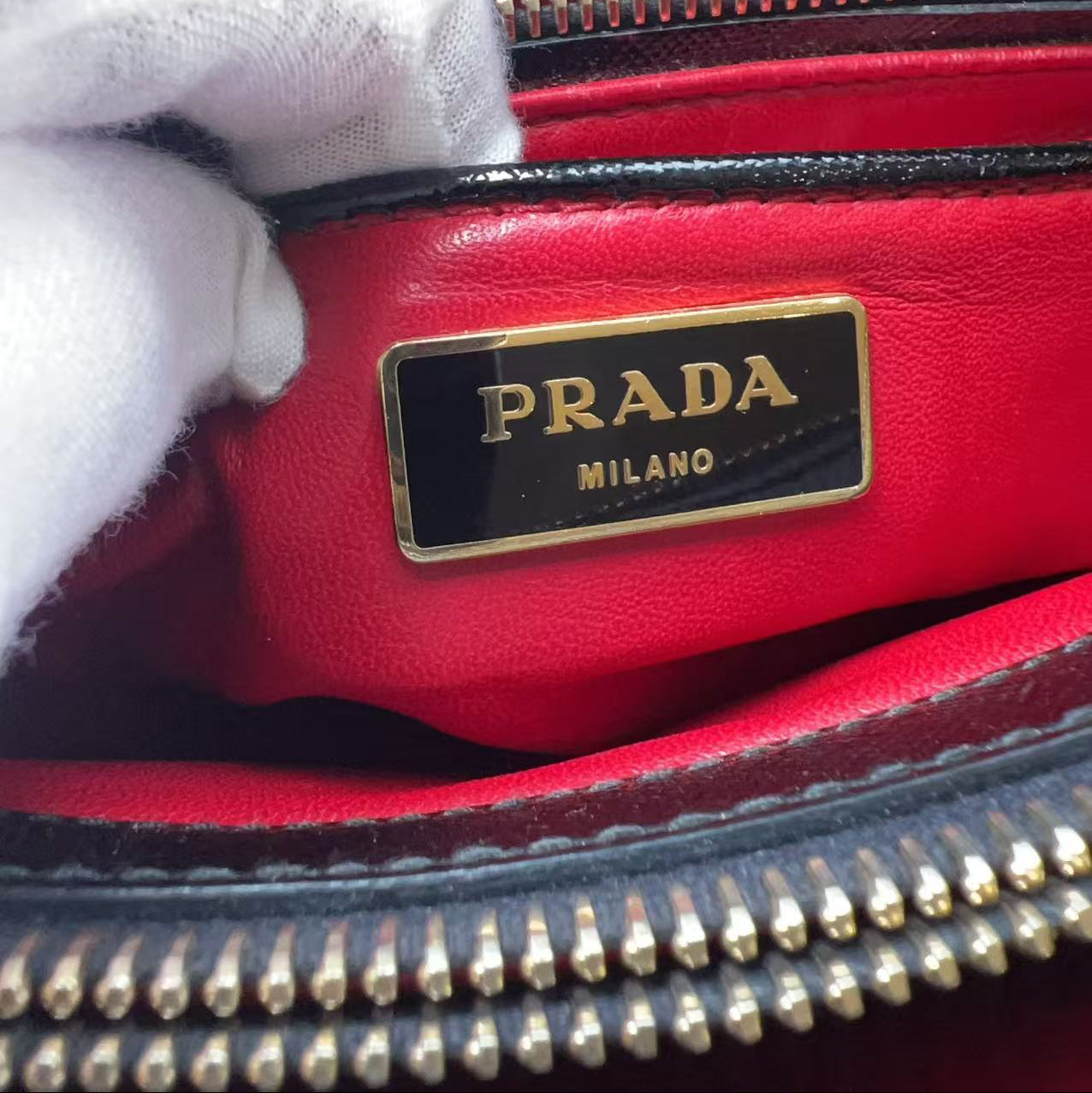 Close-up of a Prada handbag interior showing a red leather lining, gold-tone zipper, and a black plaque with gold lettering that reads PRADA MILANO. A white gloved hand is touching the bag.
