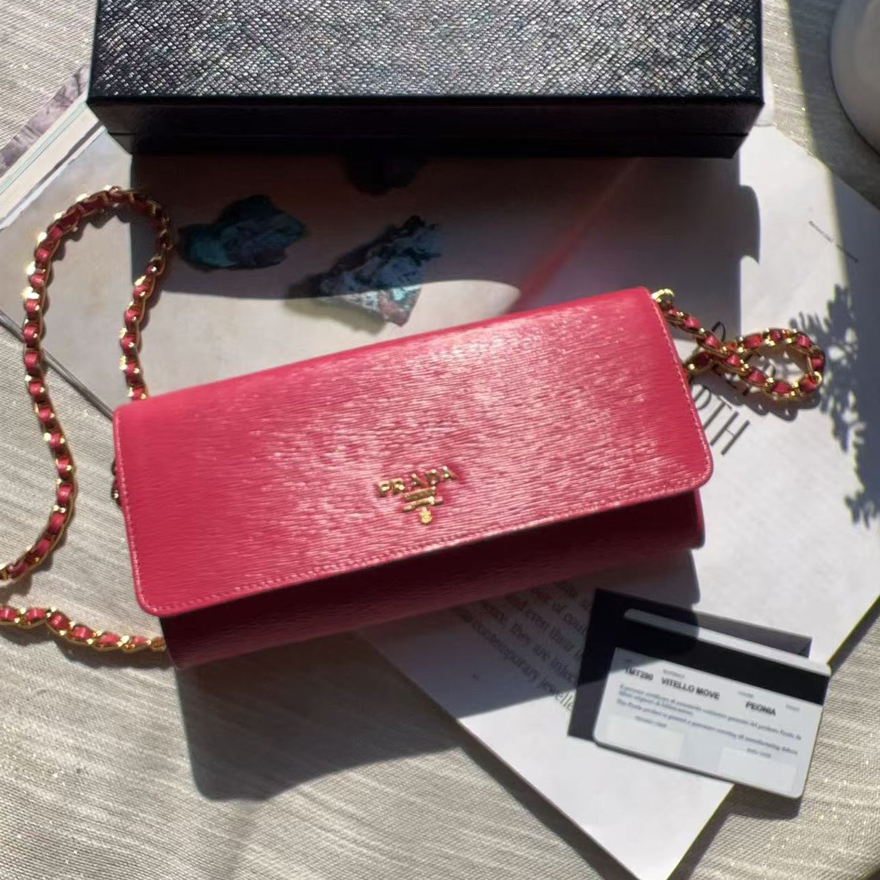 A Prada pink leather wallet or card holder with a gold logo clasp, shown next to its black textured Prada box and a red-and-gold chain strap on a light fabric surface.