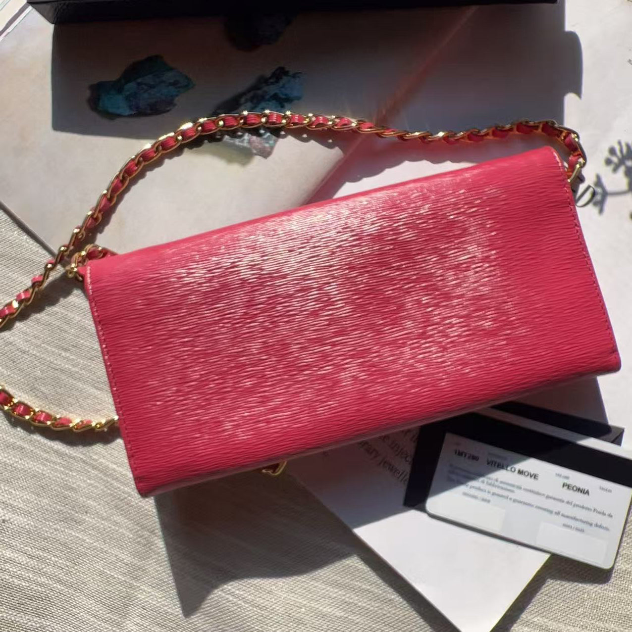 A pink textured leather clutch bag with a gold chain strap, displayed on a light-colored fabric surface alongside its black box and cards.