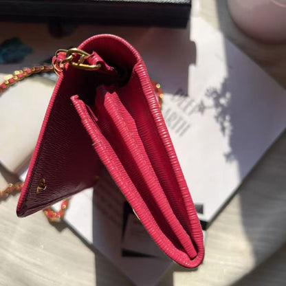 A close-up of a red leather wallet with gold-tone chain strap and hardware, photographed on a light surface with shadows and packaging nearby.