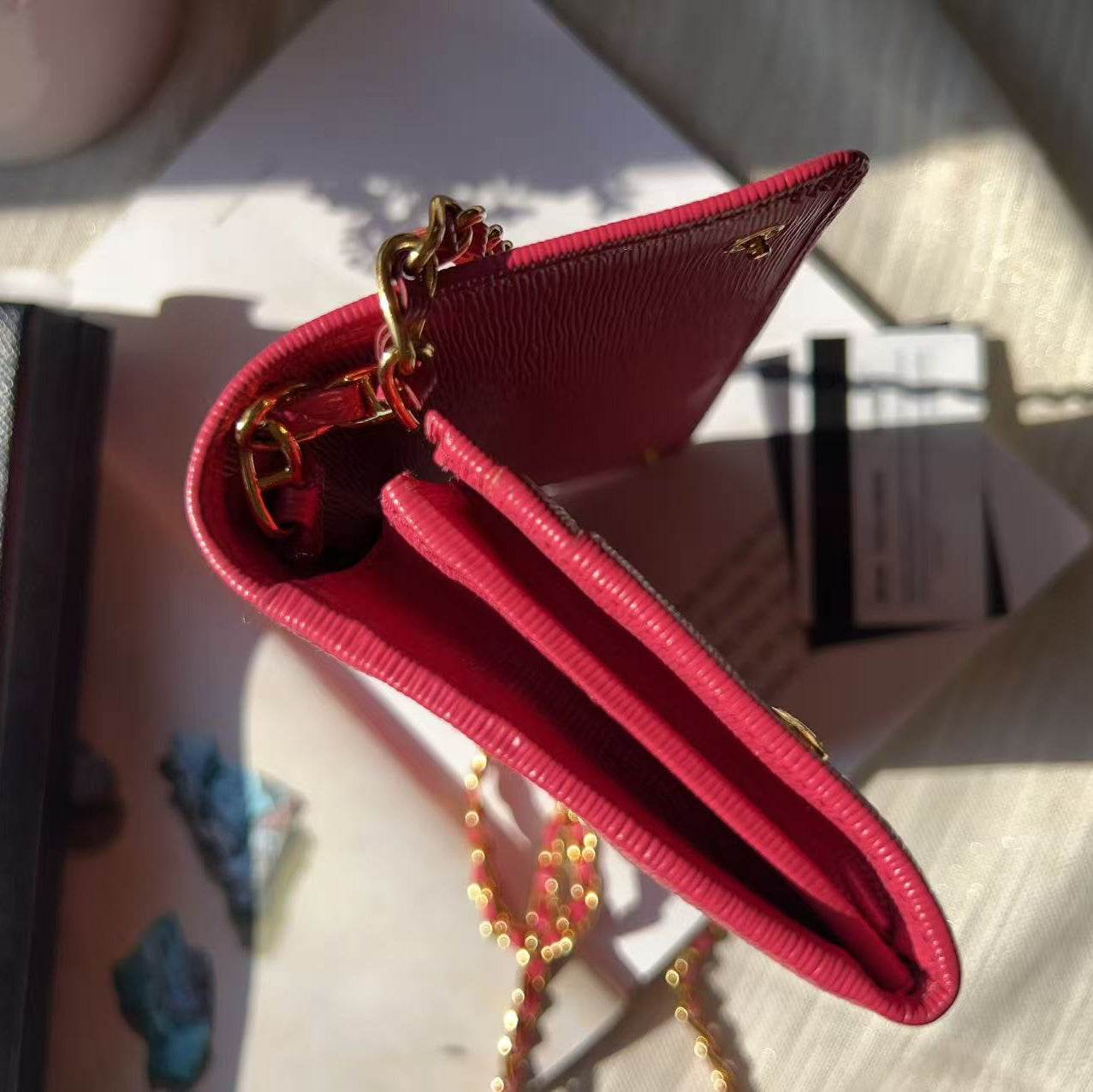 A close-up of a bright pink textured leather wallet or card holder with gold-tone chain hardware, resting on a light surface with sunlight and shadows.