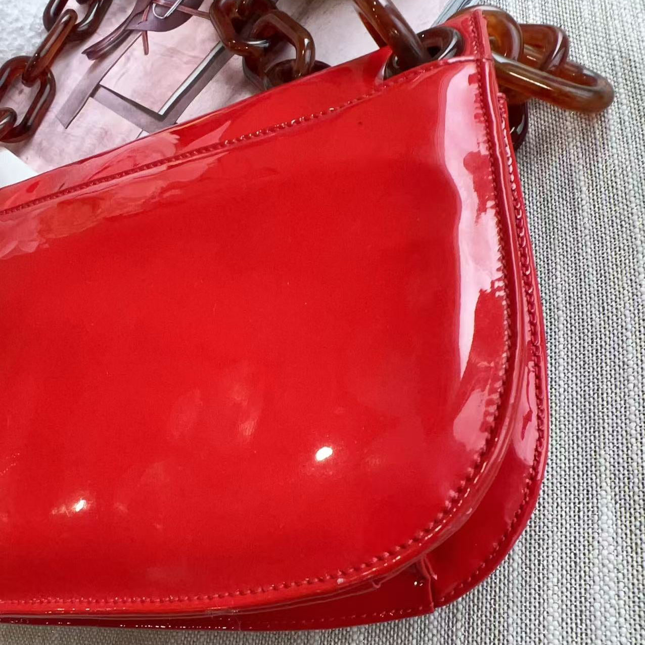 Red patent leather handbag with a brown chain handle on a textured surface.