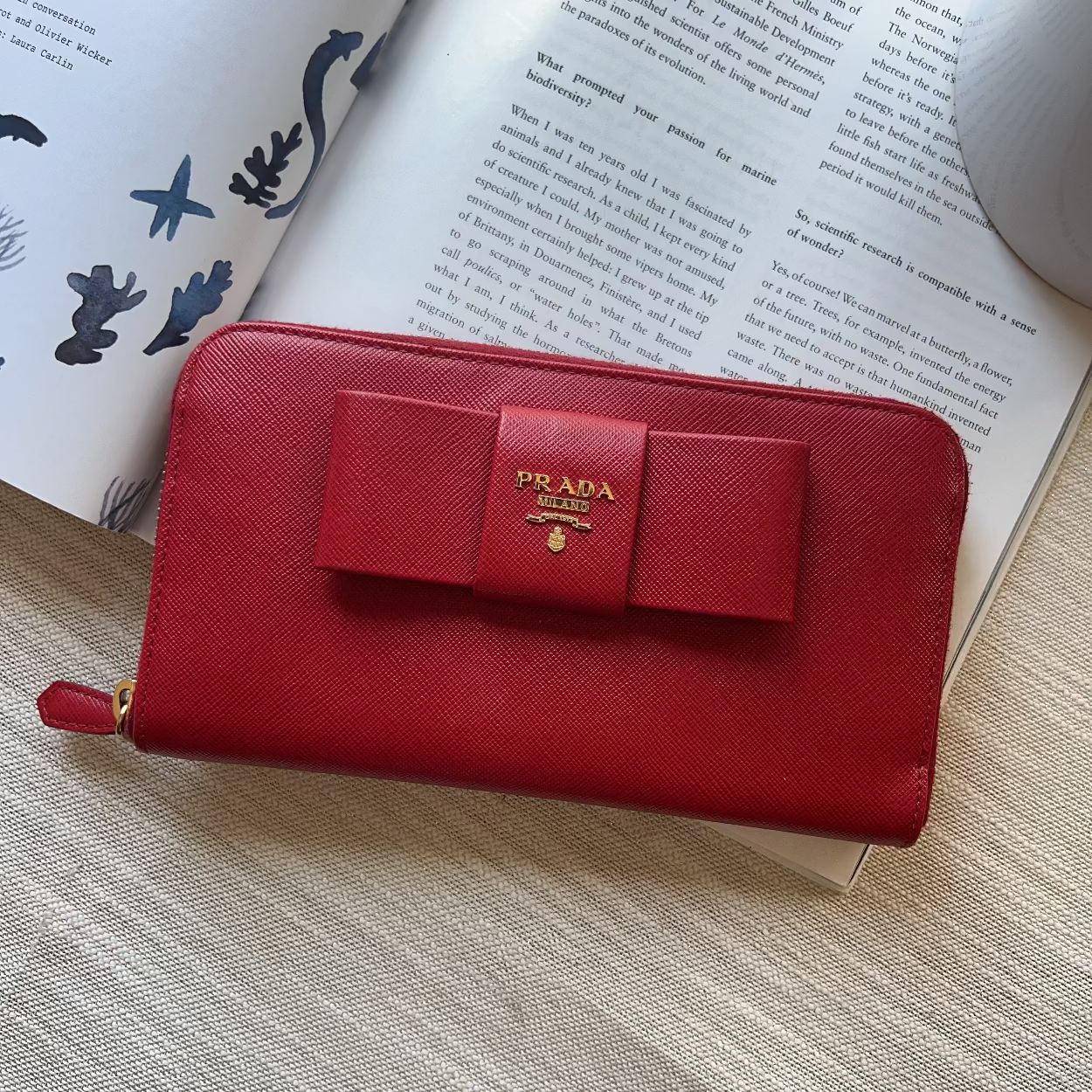 A red Prada zip-around wallet with a bow and gold logo, styled on top of an open book over a textured beige fabric surface.