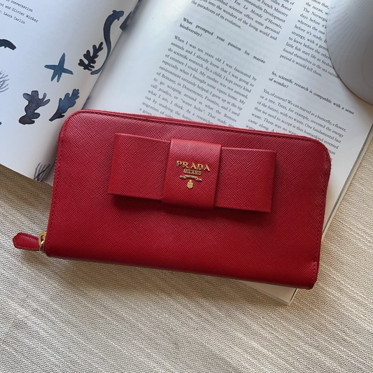 A red Prada zip-around wallet with a bow and gold logo, styled on top of an open book over a textured beige fabric surface.
