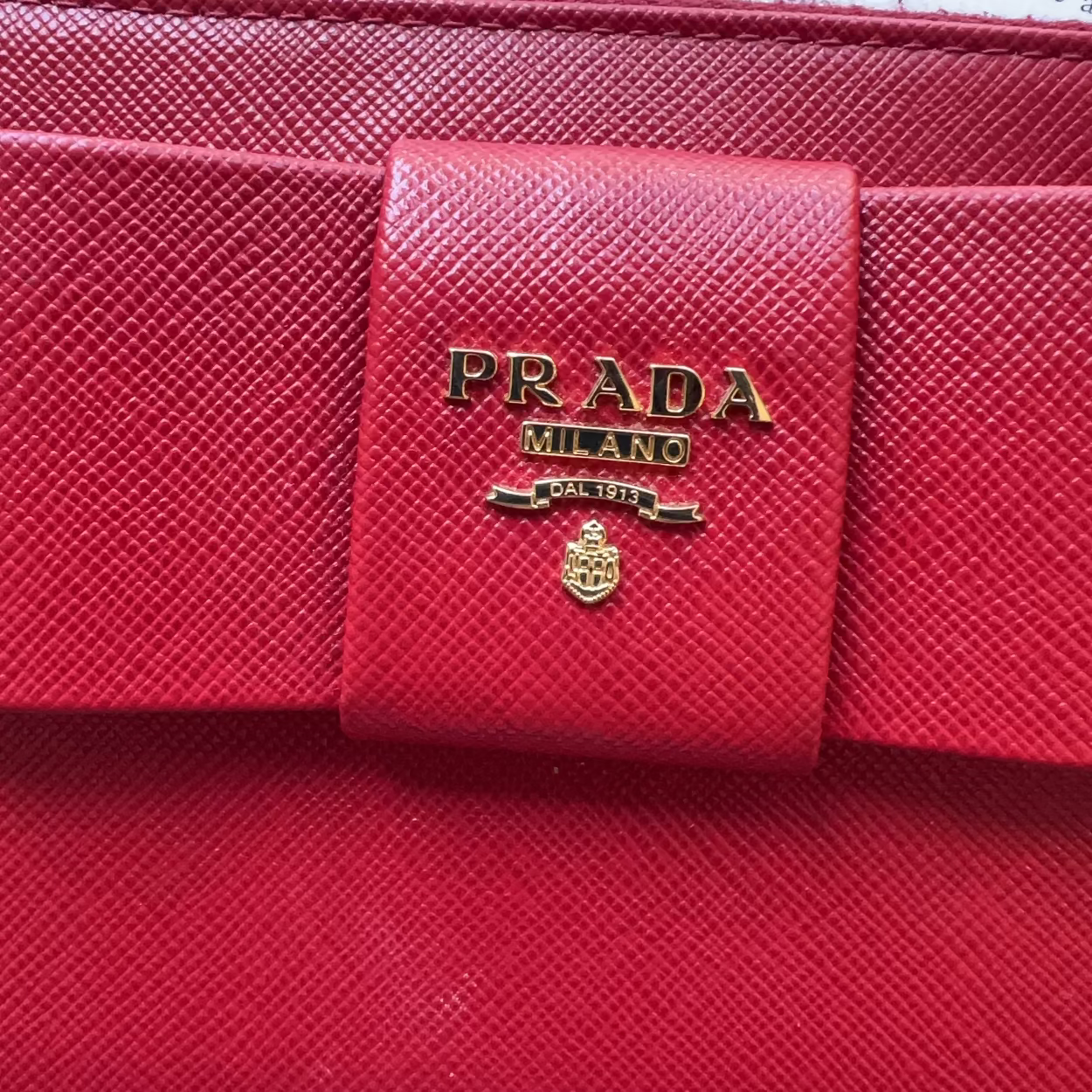 A close-up of a red Prada wallet or clutch featuring a prominent gold-tone 'PRADA MILANO' logo plaque on the front flap, set against a background of printed book pages.