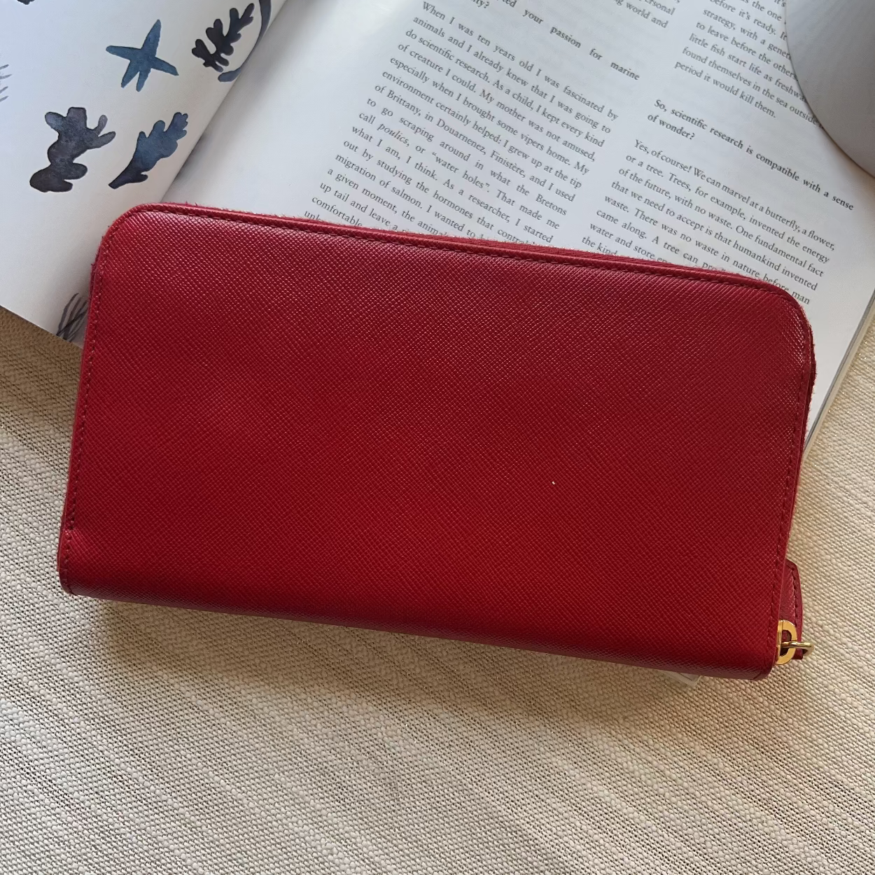 A red textured leather zip-around wallet or clutch resting on a light beige woven surface with an open book in the background.