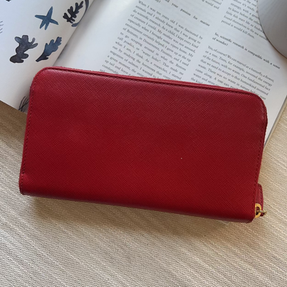 A red textured leather zip-around wallet or clutch resting on a light beige woven surface with an open book in the background.