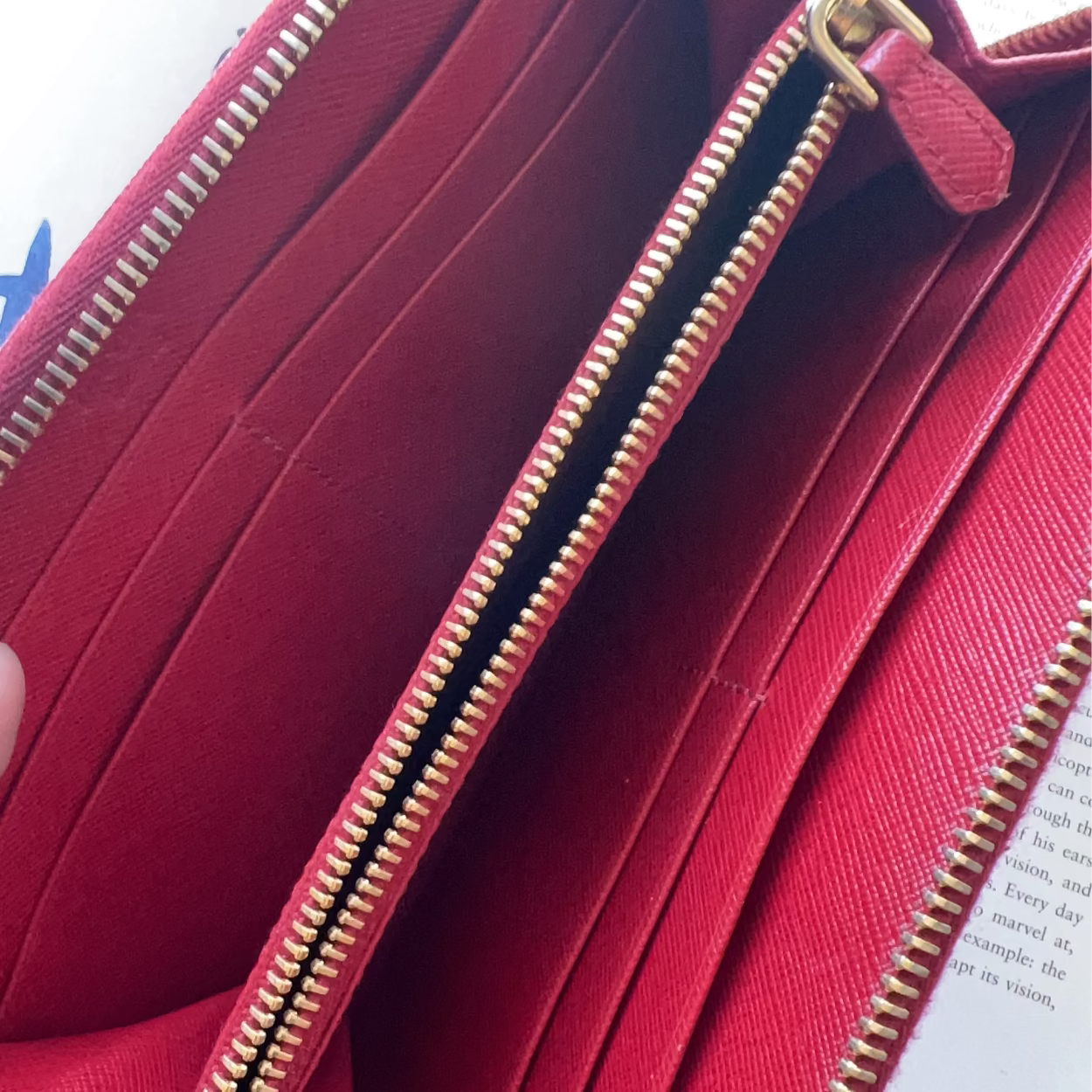 A close-up of a red leather wallet being held open by fingers, showing multiple card slots, a zip compartment, and gold-tone zippers.