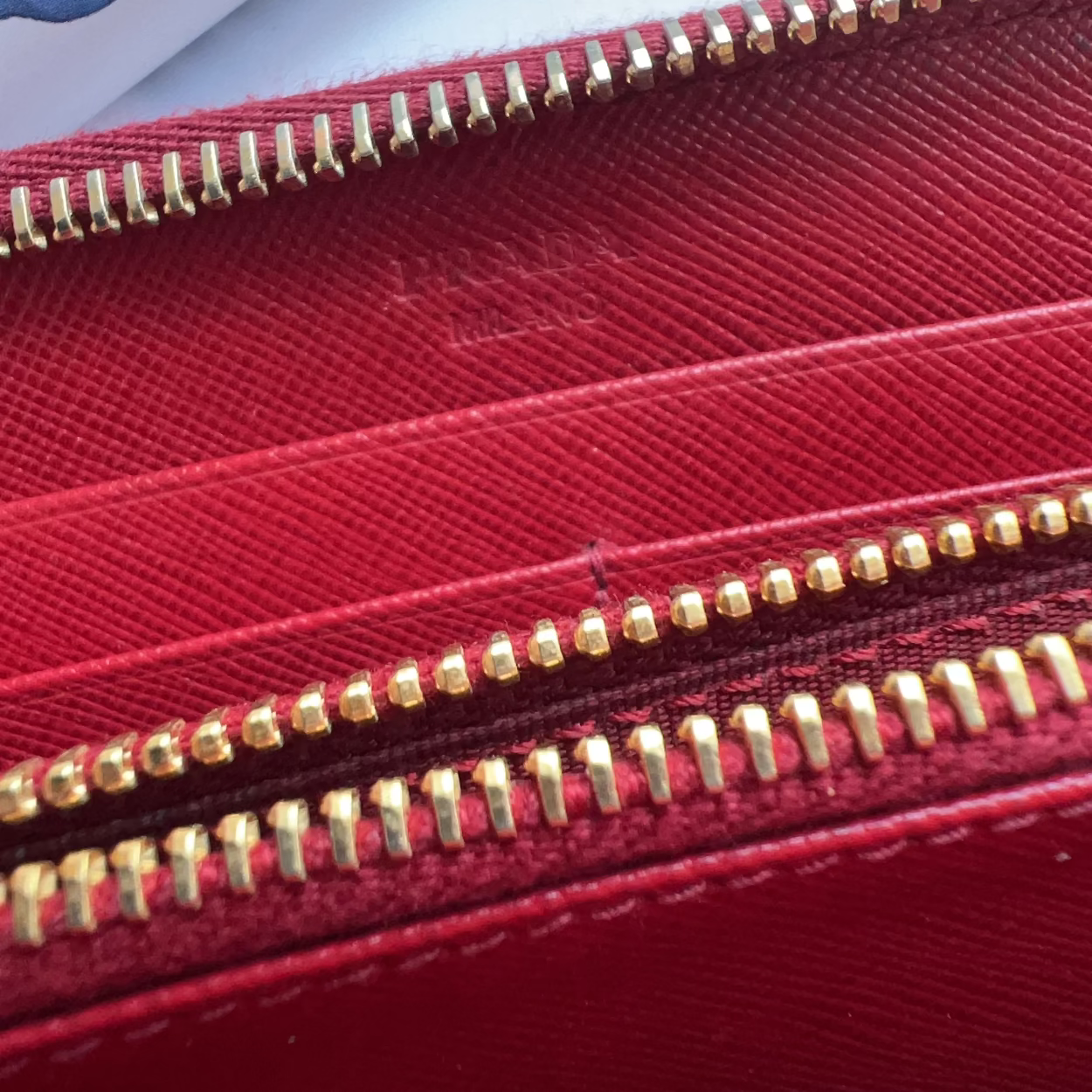 A close-up of a red textured wallet or pouch with gold-tone zippers, showing a faint embossed brand name on the interior.