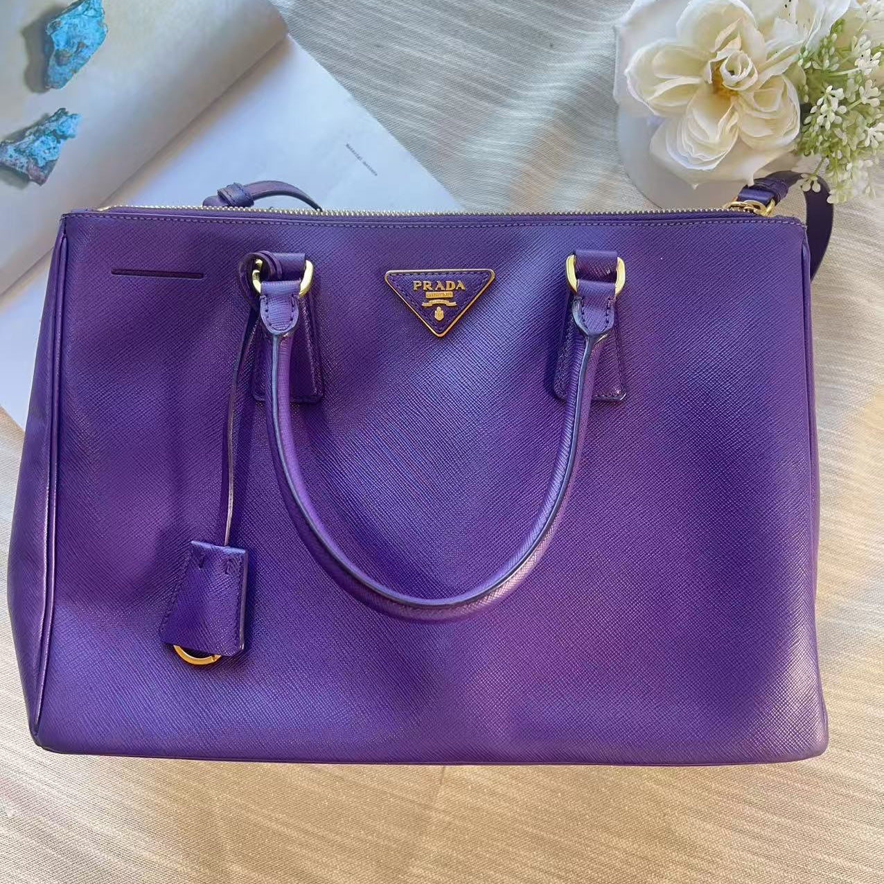 A purple Prada shoulder bag with gold-tone hardware, displayed on a light wood surface with decorative flowers and a book in the background.
