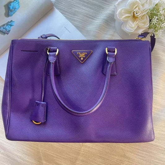 A purple Prada shoulder bag with gold-tone hardware, displayed on a light wood surface with decorative flowers and a book in the background.