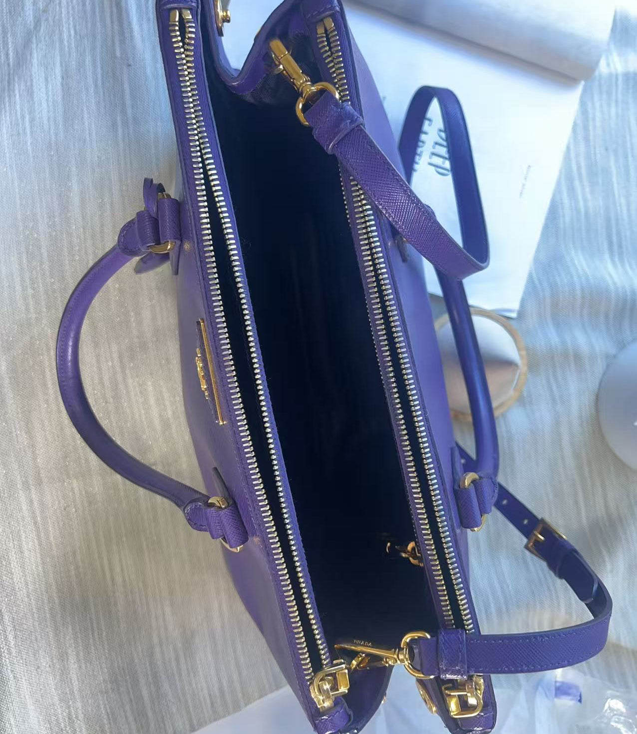 A purple leather handbag shown open from above, with gold-tone zippers, handles, and hardware, photographed on a light fabric surface.