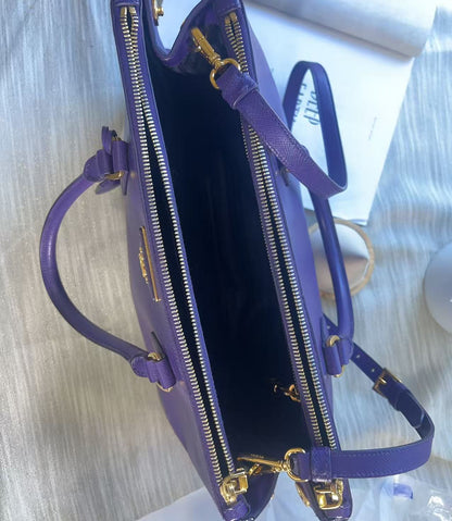 A purple leather handbag shown open from above, with gold-tone zippers, handles, and hardware, photographed on a light fabric surface.