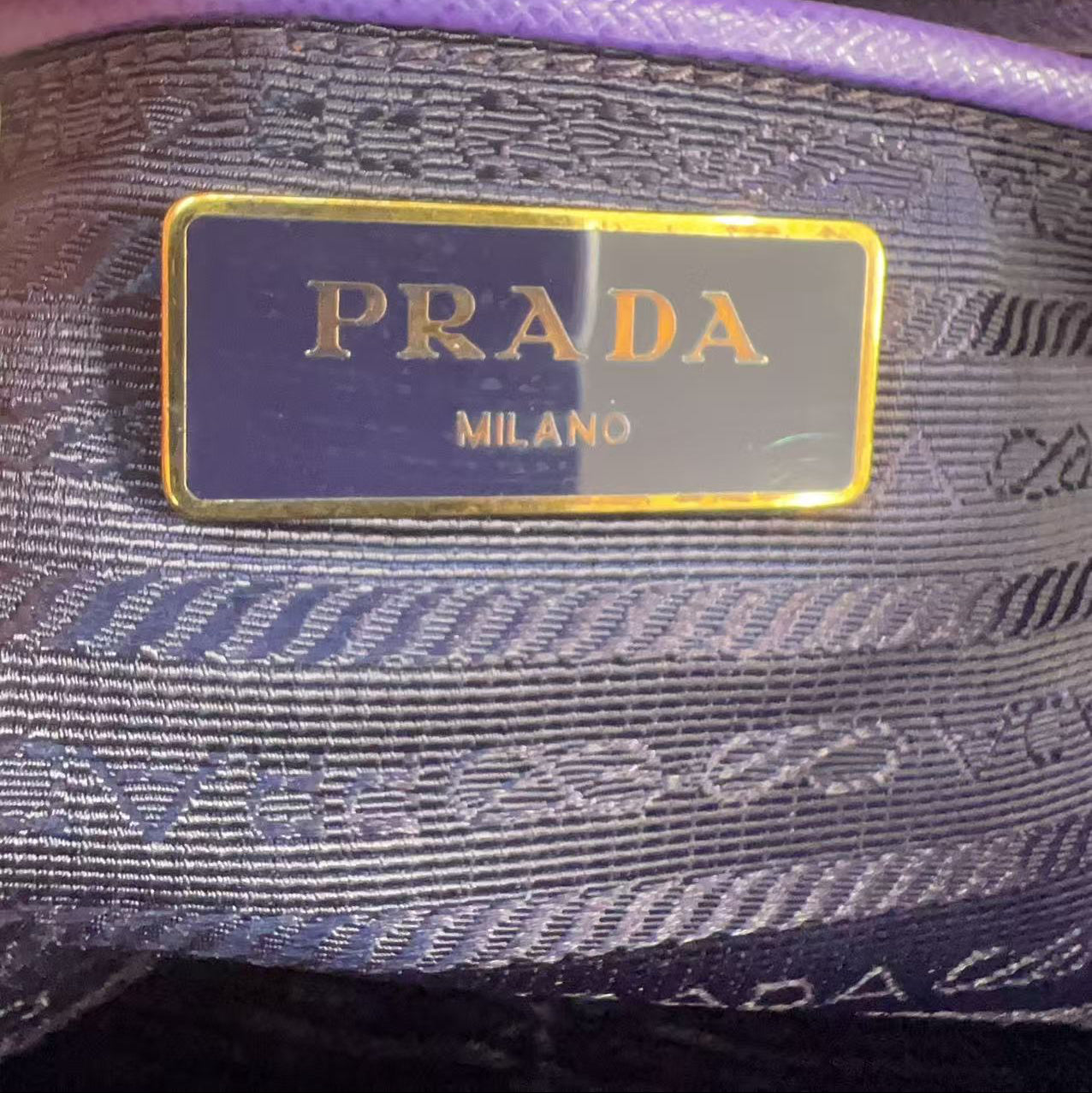 Close-up of a Prada Milano interior label on a purple and navy patterned fabric pouch with gold trim.