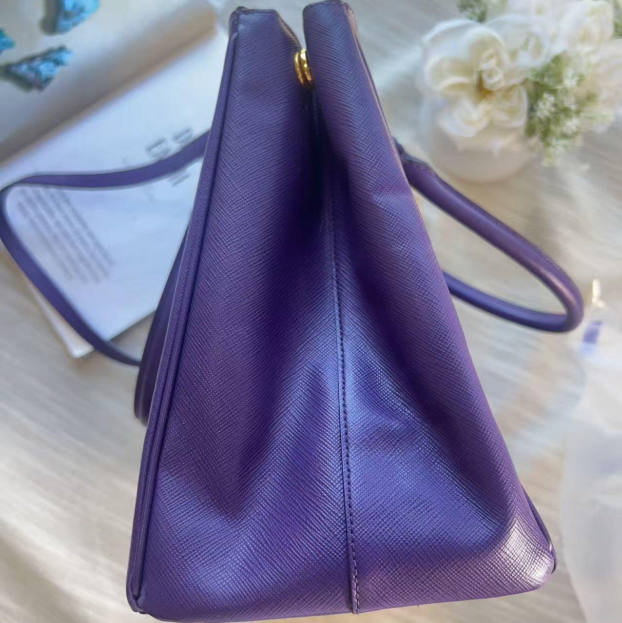 A purple textured leather handbag shown from a side angle on a light wood surface with a book and white flowers in the background.