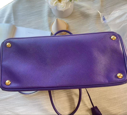 A purple textured leather handbag shown from the bottom, featuring gold-tone metal studs on the corners and a dark purple handle, resting on a light-colored wooden surface.