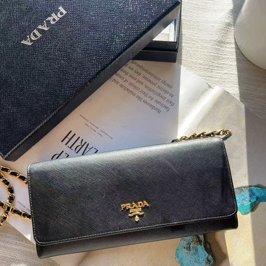 A black Prada shoulder bag with a gold-tone logo and chain strap, styled on a beige fabric surface with a blue Prada box, printed paper, and turquoise stones.
