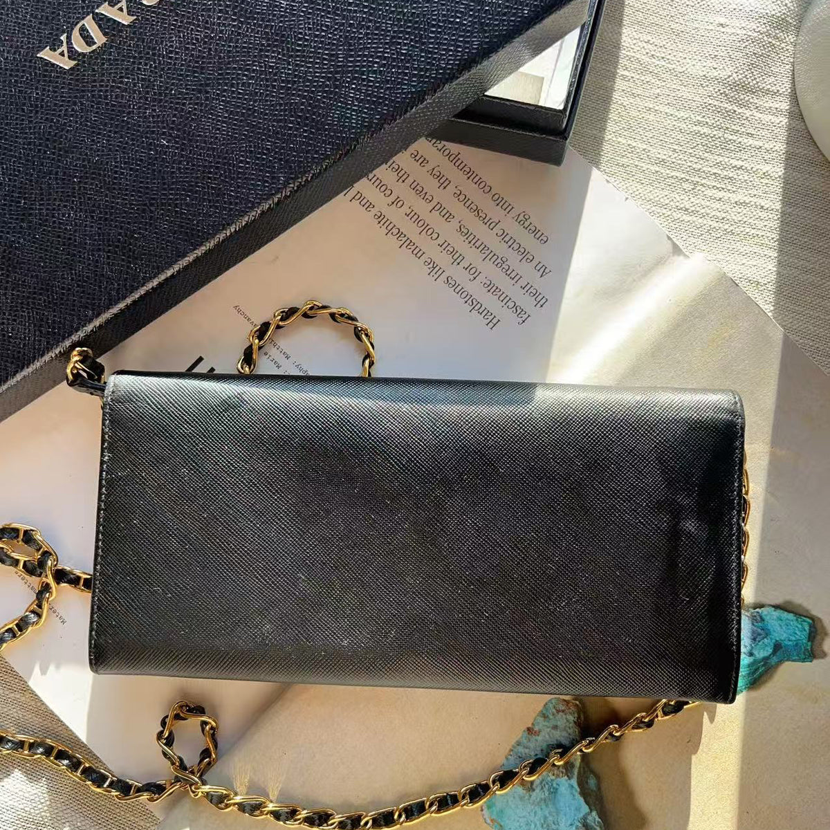 A black Prada shoulder bag with a gold chain strap, displayed on a beige surface next to its navy blue textured Prada box and turquoise stones.