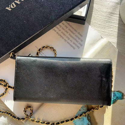 A black Prada shoulder bag with a gold chain strap, displayed on a beige surface next to its navy blue textured Prada box and turquoise stones.