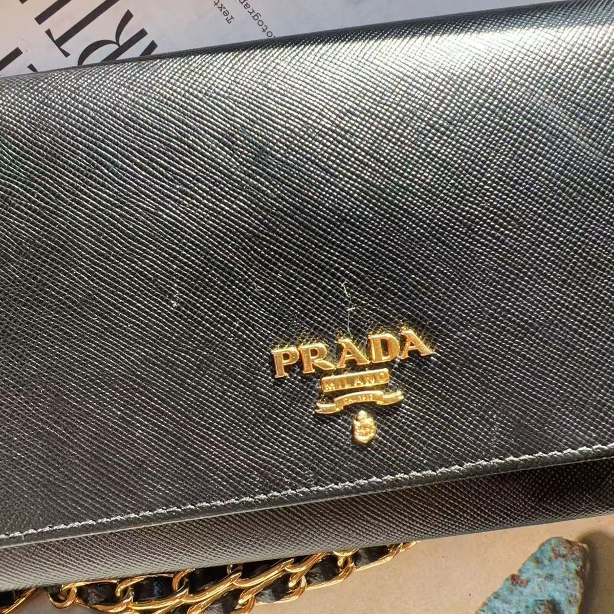 A close-up of a Prada wallet or clutch in textured black leather with a gold-tone Prada logo plaque on the front, resting on a beige surface with a chain strap partially visible.