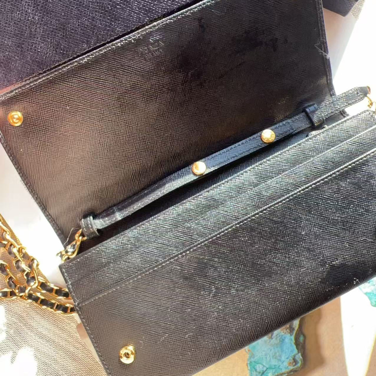 A close-up of a black textured leather handbag with gold-tone hardware, showing the interior flap, strap attachment, and part of a chain strap.