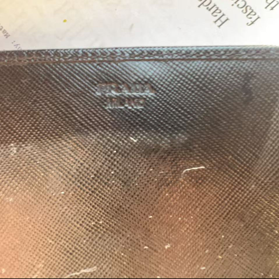 A close-up of a dark brown textured leather or canvas item with an embossed Prada logo, resting on a piece of paper with printed text.