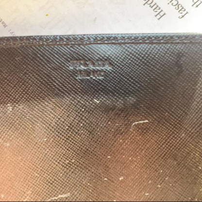 A close-up of a dark brown textured leather or canvas item with an embossed Prada logo, resting on a piece of paper with printed text.