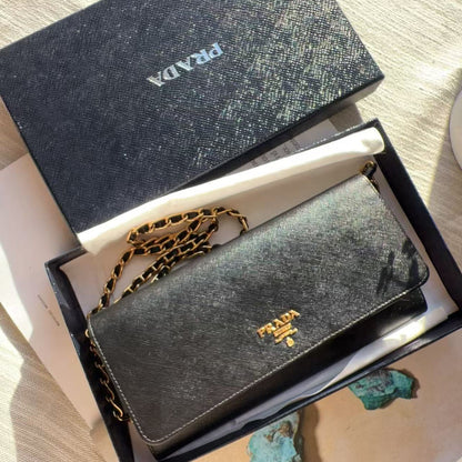 A Prada wallet in textured black leather with a gold-tone chain strap, presented in its black box alongside the branded outer lid on a light fabric surface.