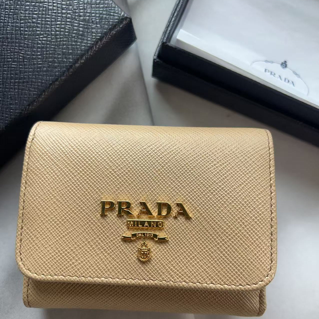 A beige Prada Saffiano wallet with gold-tone logo hardware, displayed on a white surface with its black textured presentation box and white insert visible in the background.