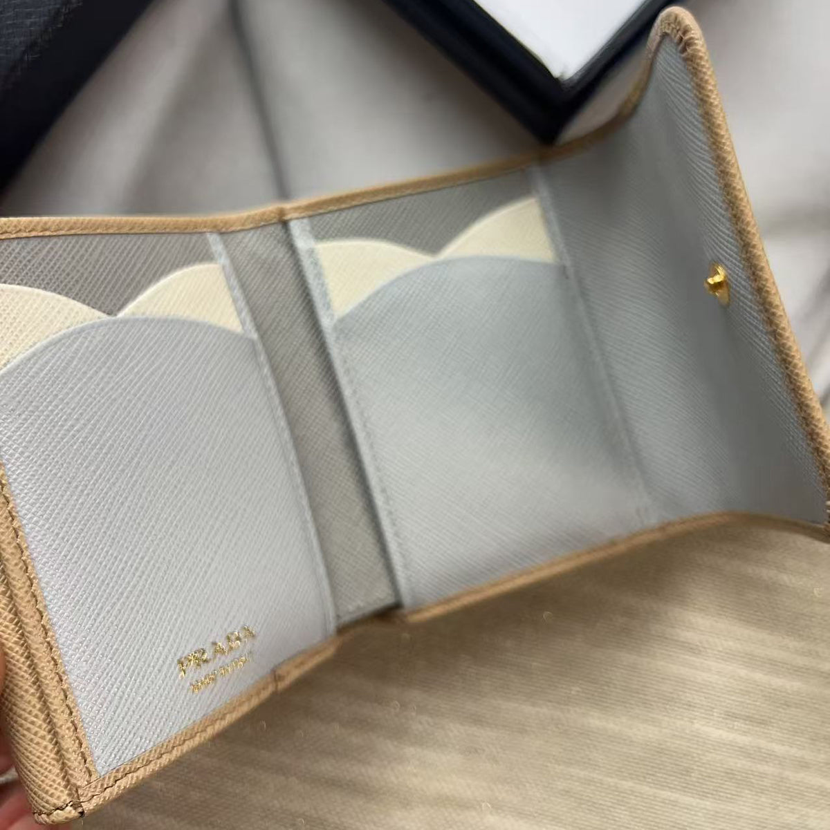 An open Prada wallet in light gray saffiano leather with beige trim, shown on a light wood surface with the brand's packaging in the background.
