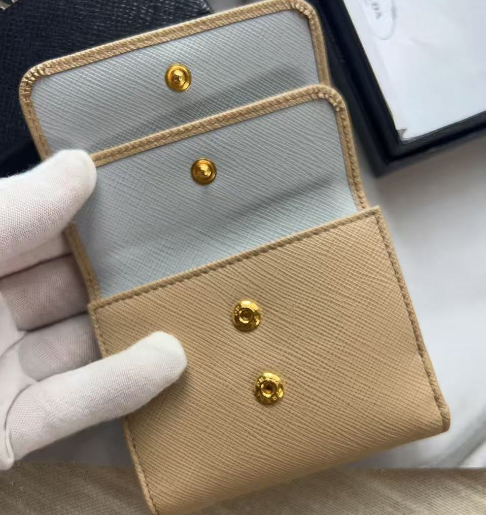 A close-up of a small leather card holder or wallet with a tan exterior, light gray interior, and three gold-tone snap buttons, held by a white-gloved hand.