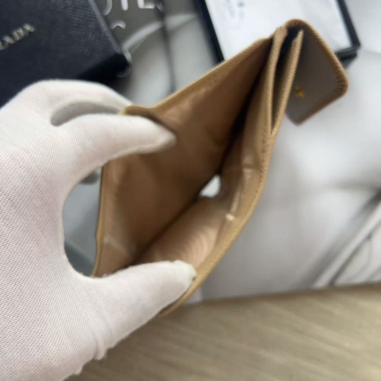 A tan leather card holder or wallet being held open by a white-gloved hand, with a branded black box and white surface in the background.