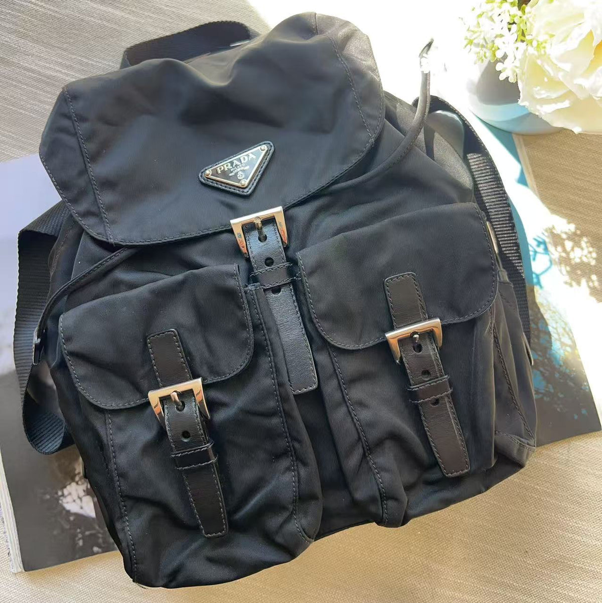 A black nylon Prada shoulder bag with multiple front pockets, brown leather straps, and gold-tone buckles, resting on a light wood surface.