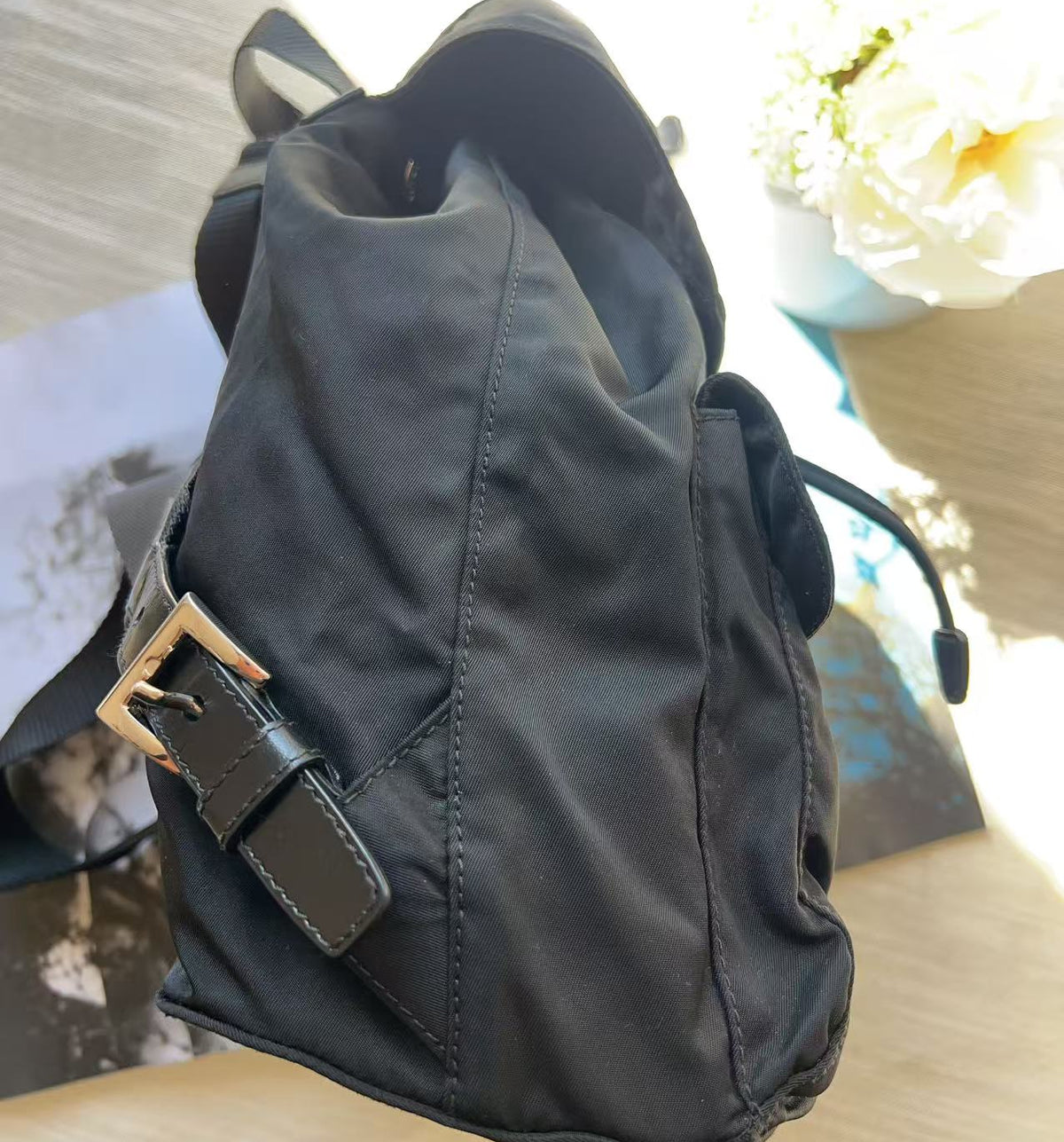 A black nylon backpack with a gold-tone buckle strap, resting on a light wooden surface next to a white flower in a vase.