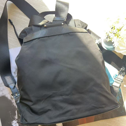 A black nylon backpack with a top handle and adjustable shoulder straps, resting on a light-colored wooden surface next to a magazine and a small plant.